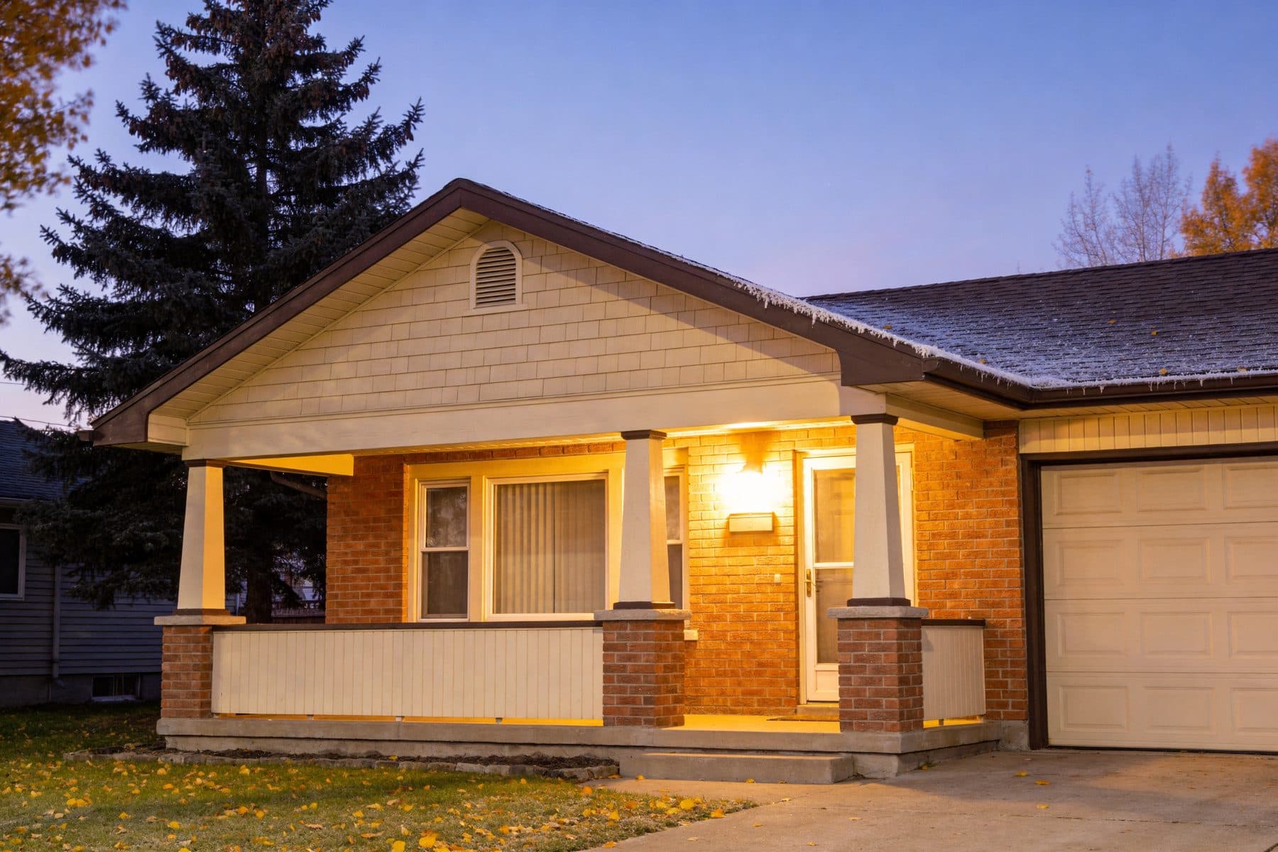 A brick bungalow on an Edmonton street at dusk in early fall, porch light on